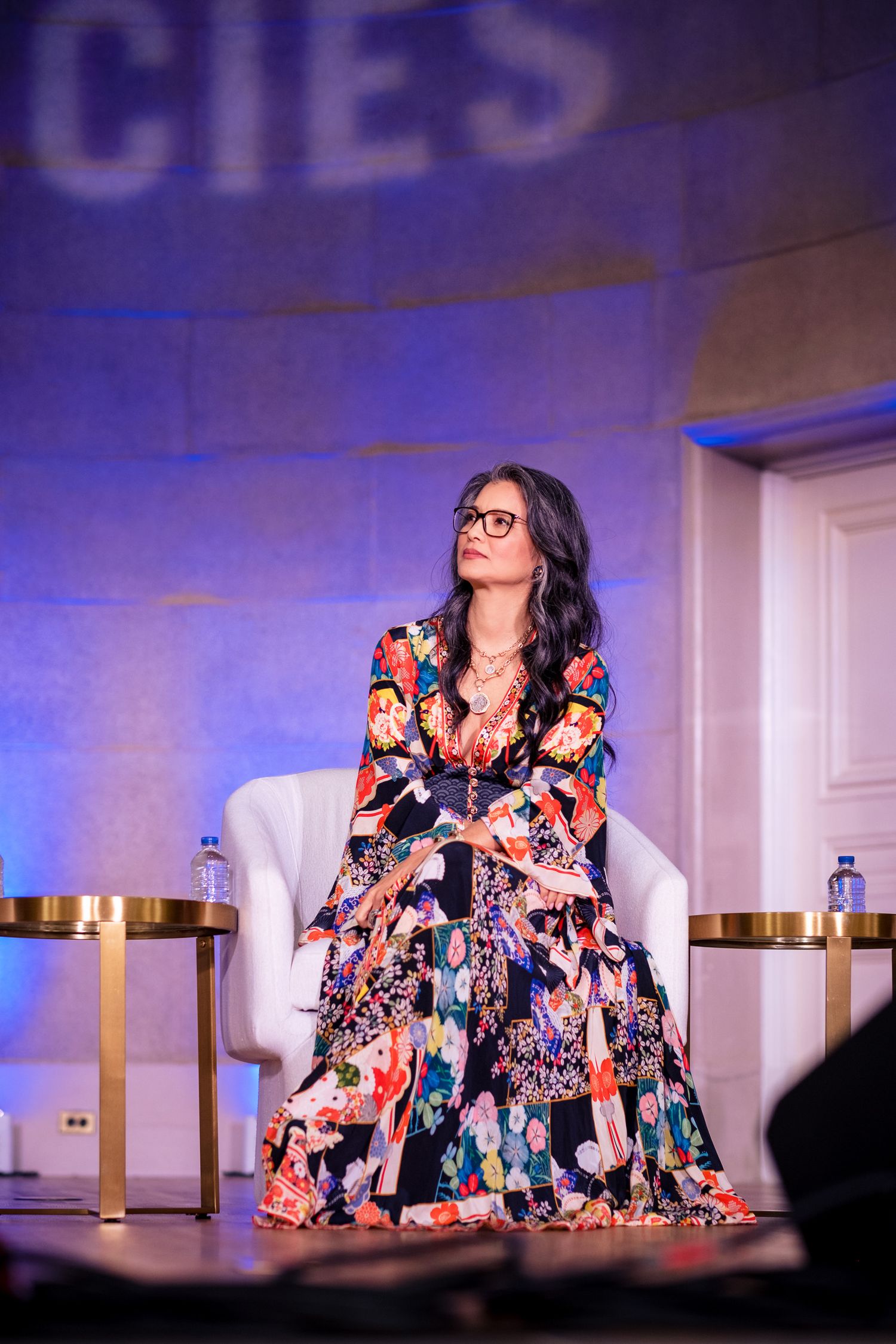 Woman in colorful floral maxi dress and glasses seated in white chair on stage at conference event.
