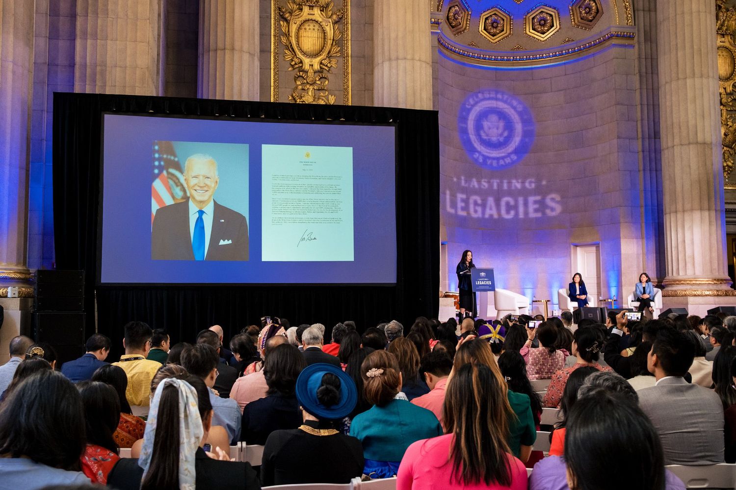 A large audience watches a projected image of President Biden at a Lasting Legacies award ceremony in a grand hall.