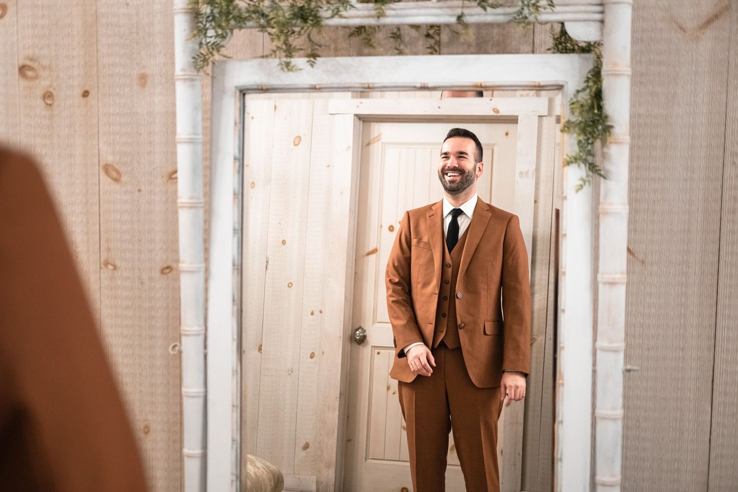 Bearded groom in elegant brown suit smiling confidently in rustic mirror reflection.