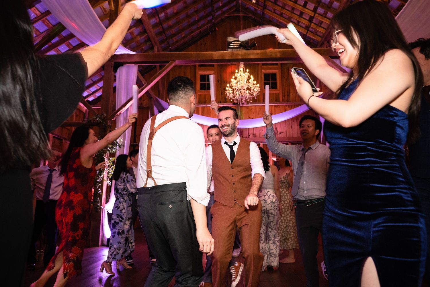 Joyful wedding guests dance together under chandelier lights in a rustic barn reception hall.