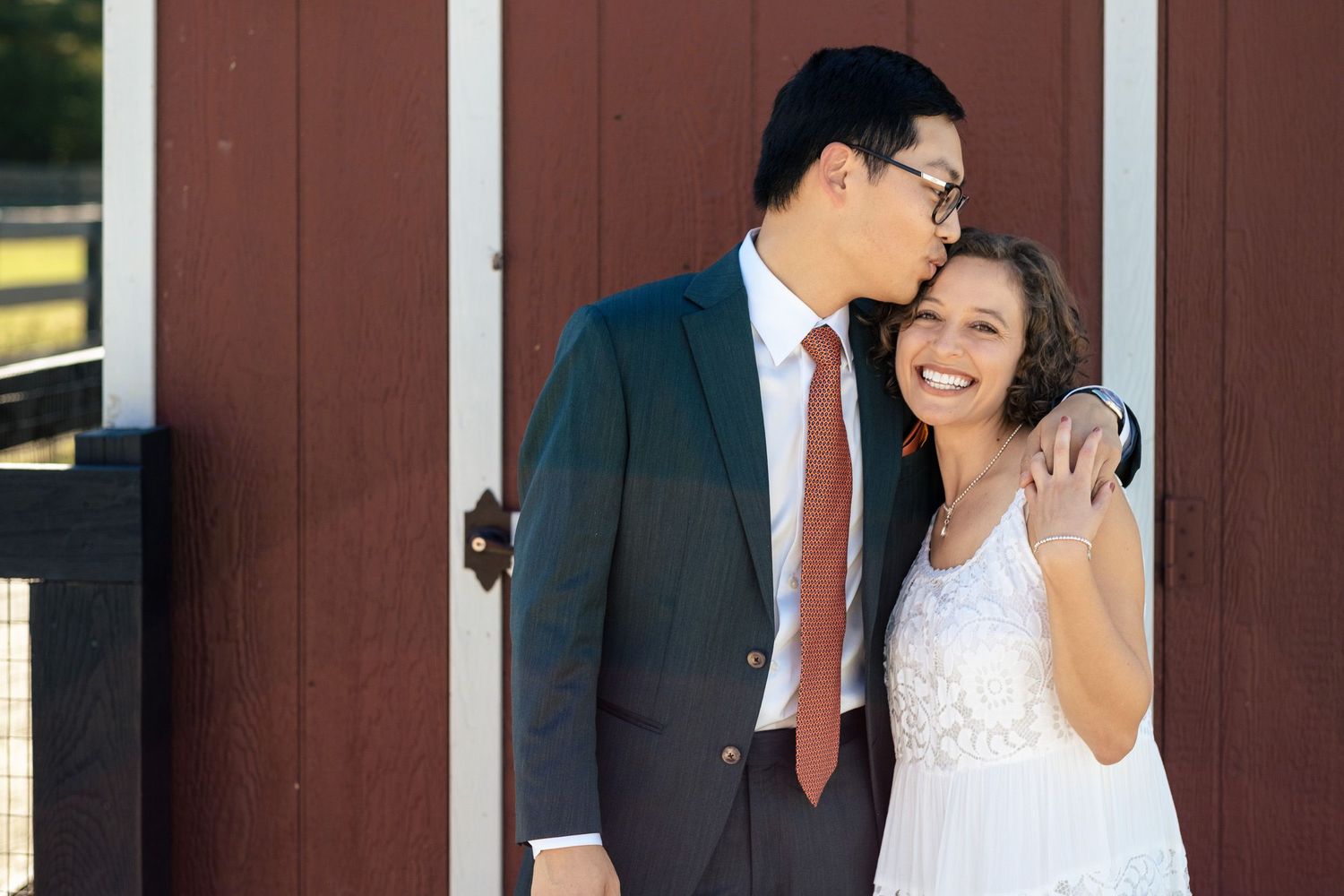 A groom tenderly kisses his smiling bride on the forehead near a red barn door.