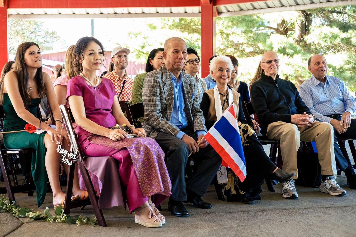 Diverse wedding guests seated outdoors, one holding a Thai flag during a wedding ceremony.