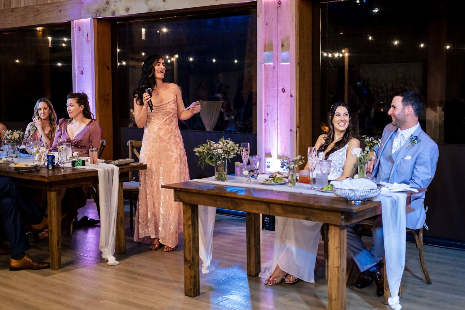 Bridesmaid giving heartfelt speech at wedding reception while couple smiles at head table.