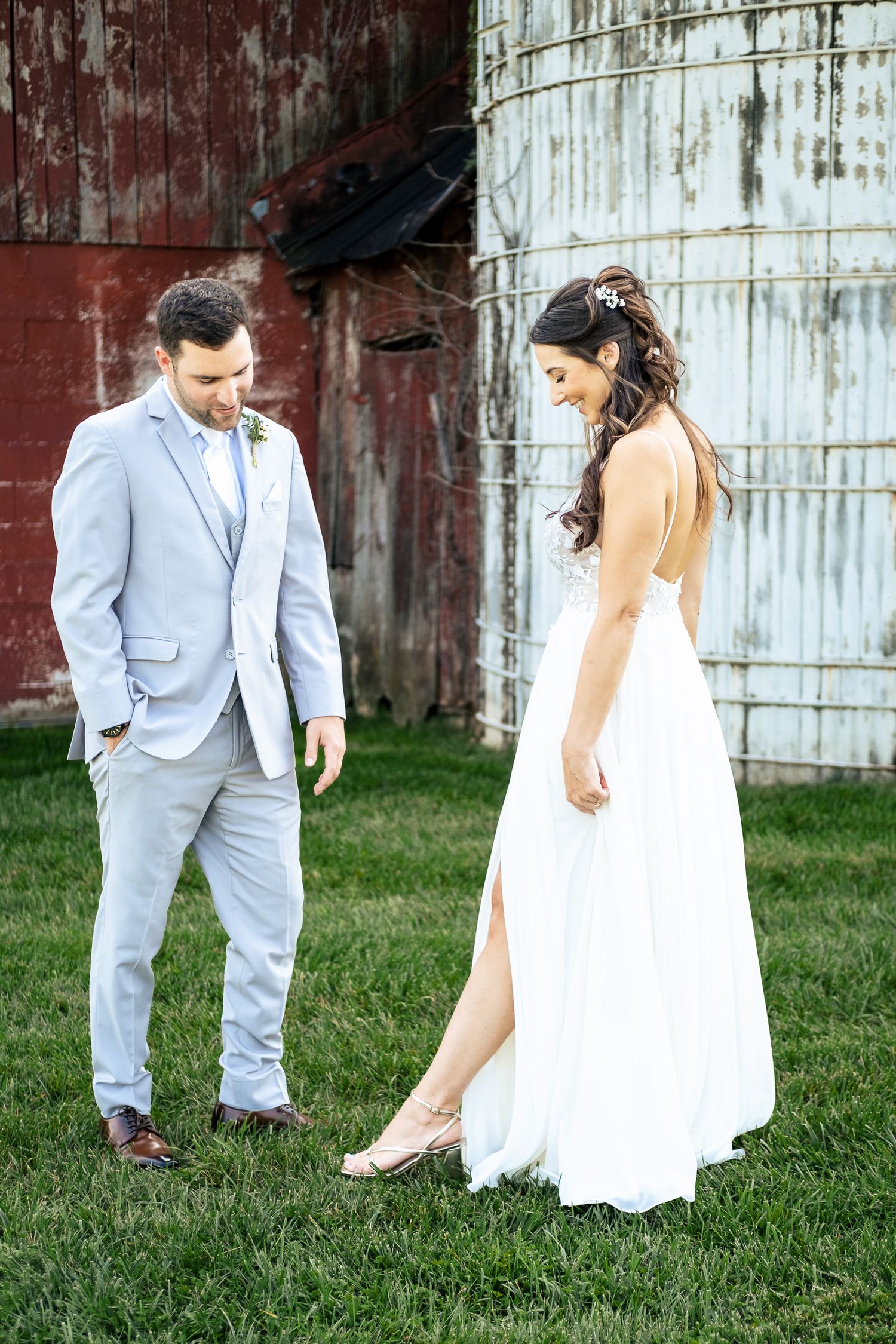Bride in white gown and groom in gray suit laugh together beside red barn and grain silo.