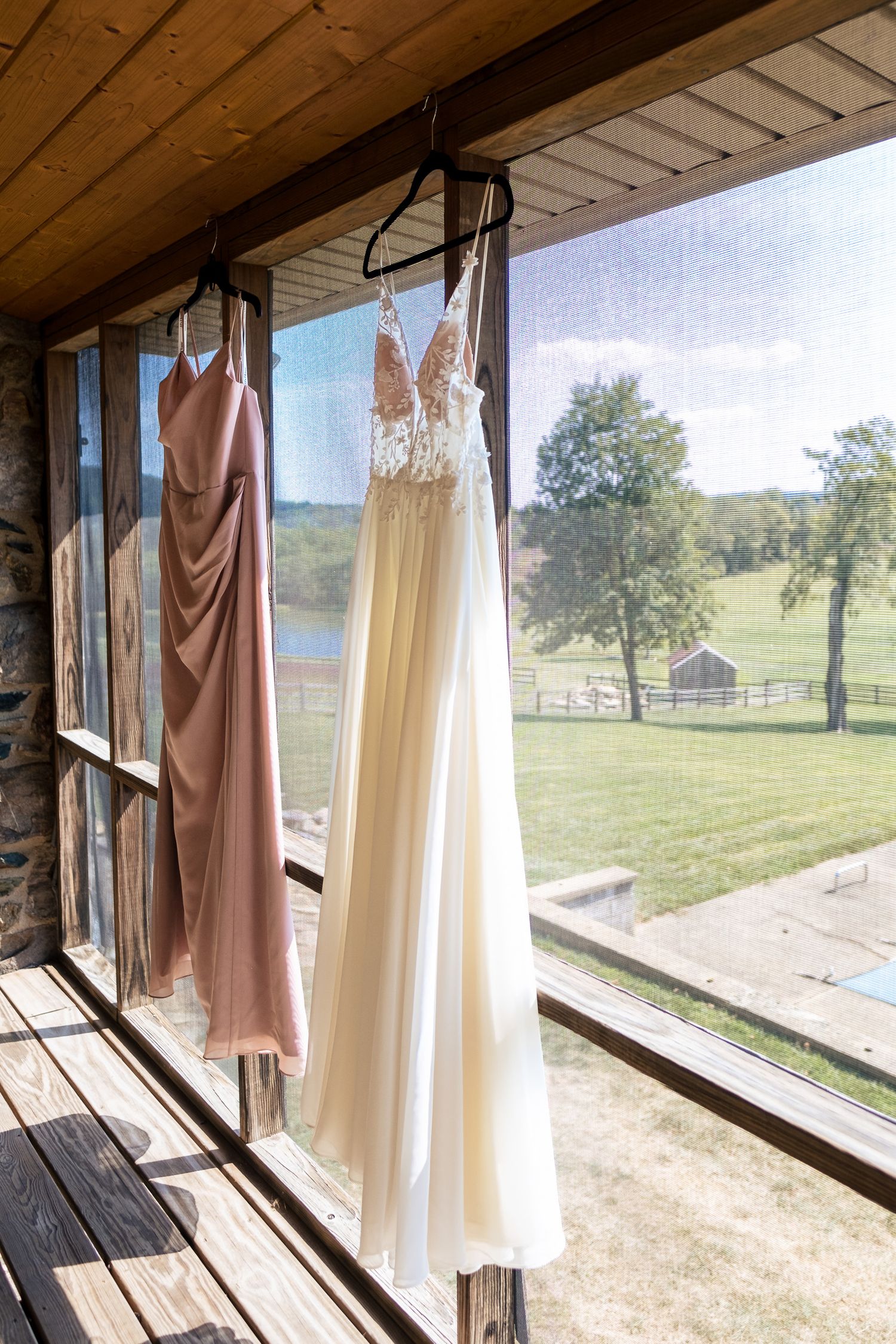 Wedding dress and bridesmaid gown hanging in a sunlit window with a scenic green countryside view.