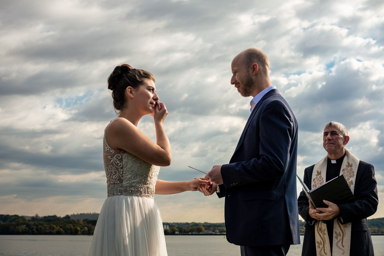 Outdoor wedding ceremony by a lake with bride wiping tears, groom holding hands, and officiant in background under cloudy sky.