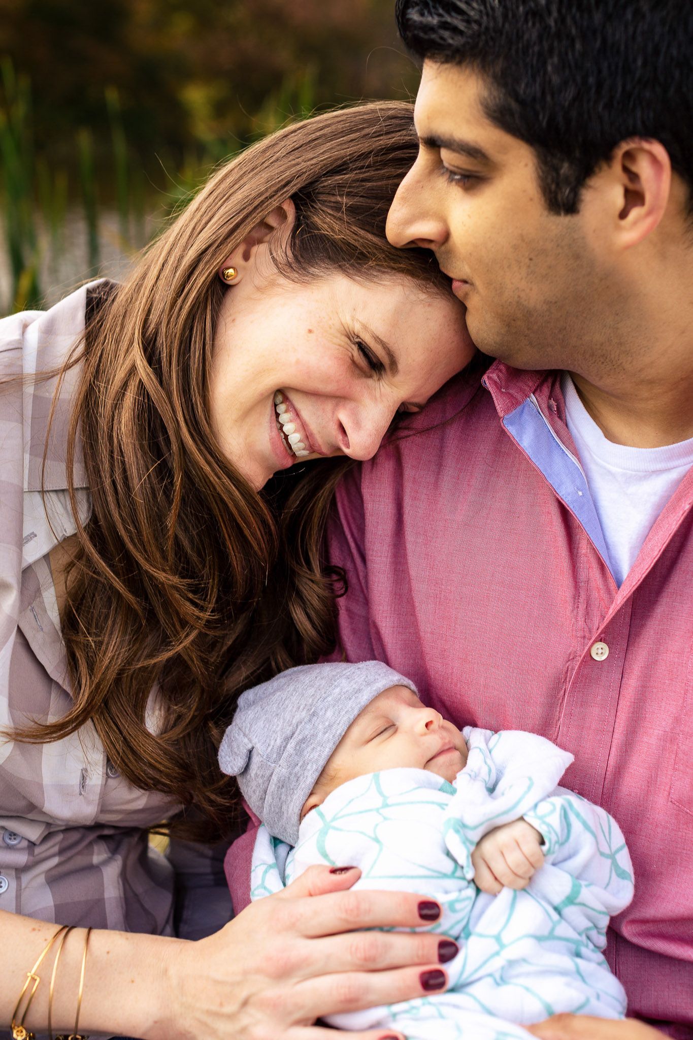 Smiling couple tenderly holding their newborn baby wrapped in a blue blanket outdoors in soft light.