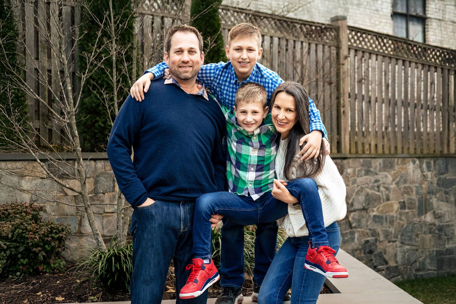 Family of four smiling together outdoors near wooden fence in casual portrait photo.