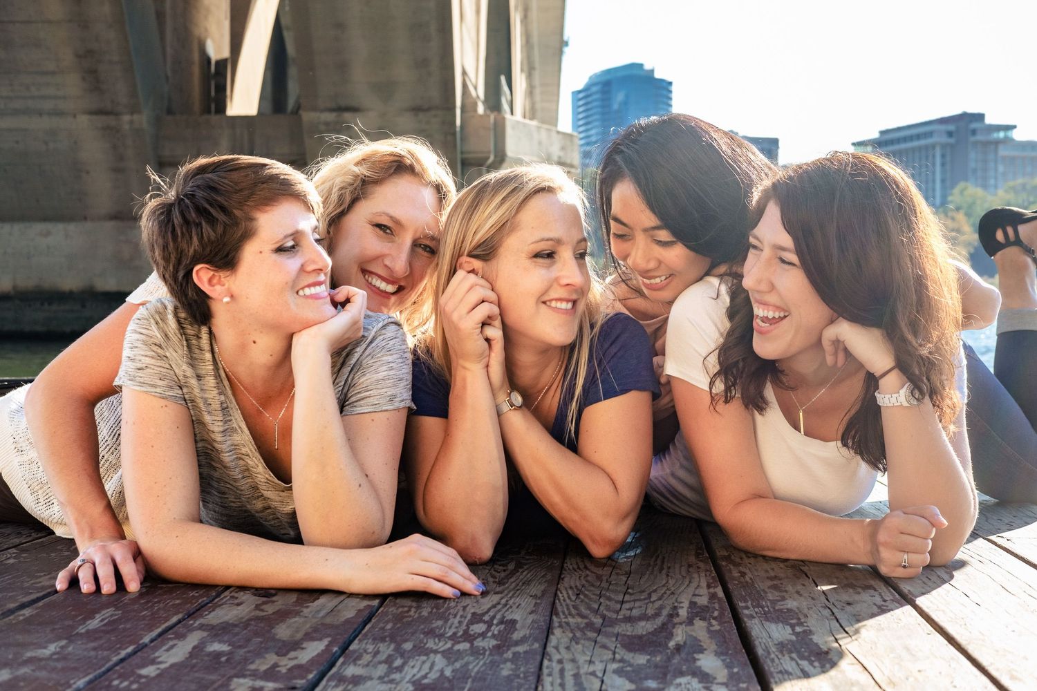 Five young women laughing and posing together on a wooden deck with city skyline behind.