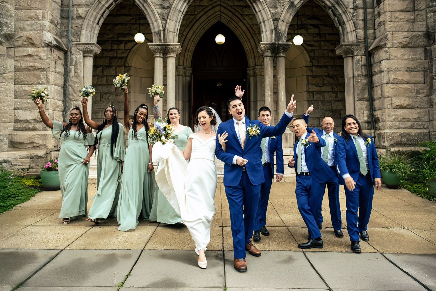 Wedding party in blue suits and mint dresses celebrate outside a grand stone church entrance.