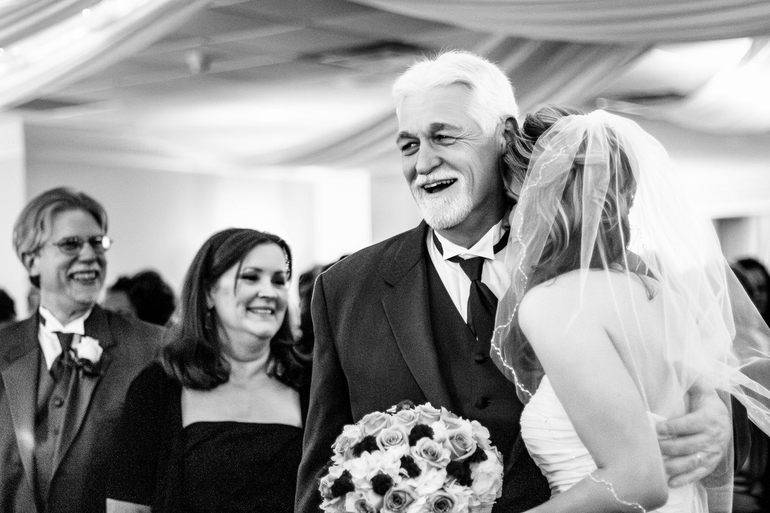 Joyful father laughs as bride hugs him at wedding reception in black and white.