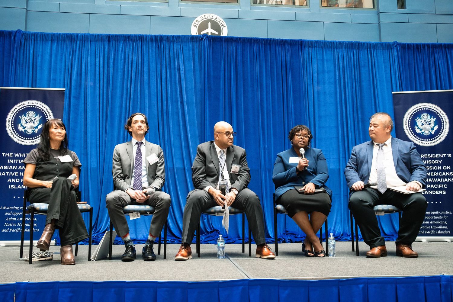 Five panelists seated on stage at a formal government conference with blue drape backdrop and official seals.