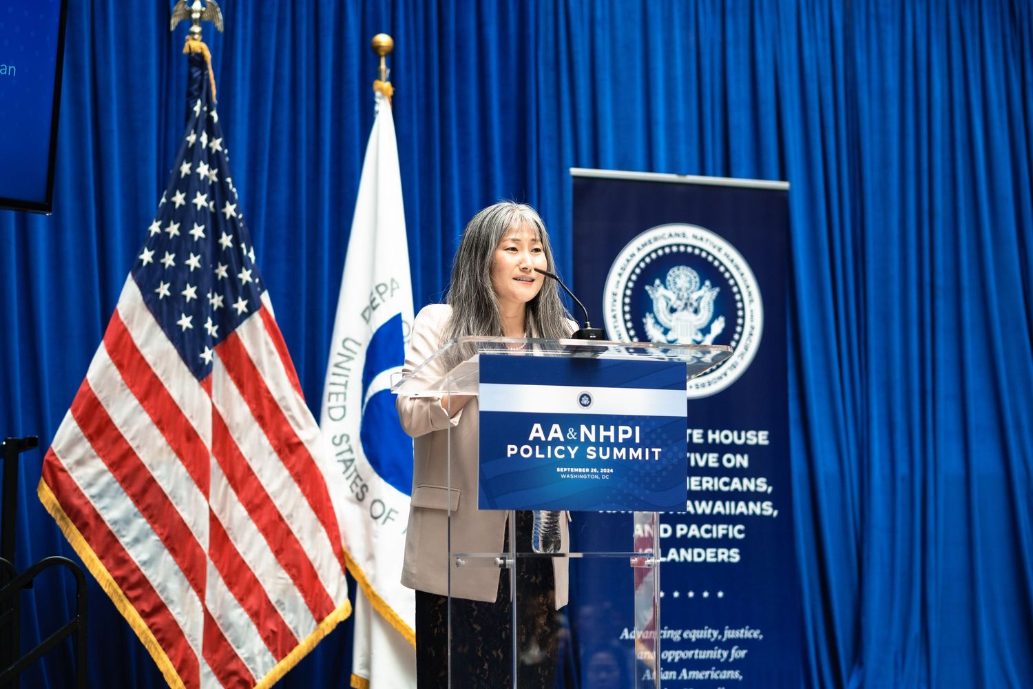 Woman speaks at podium during AA and NHPI White House Policy Summit with American flags in background.