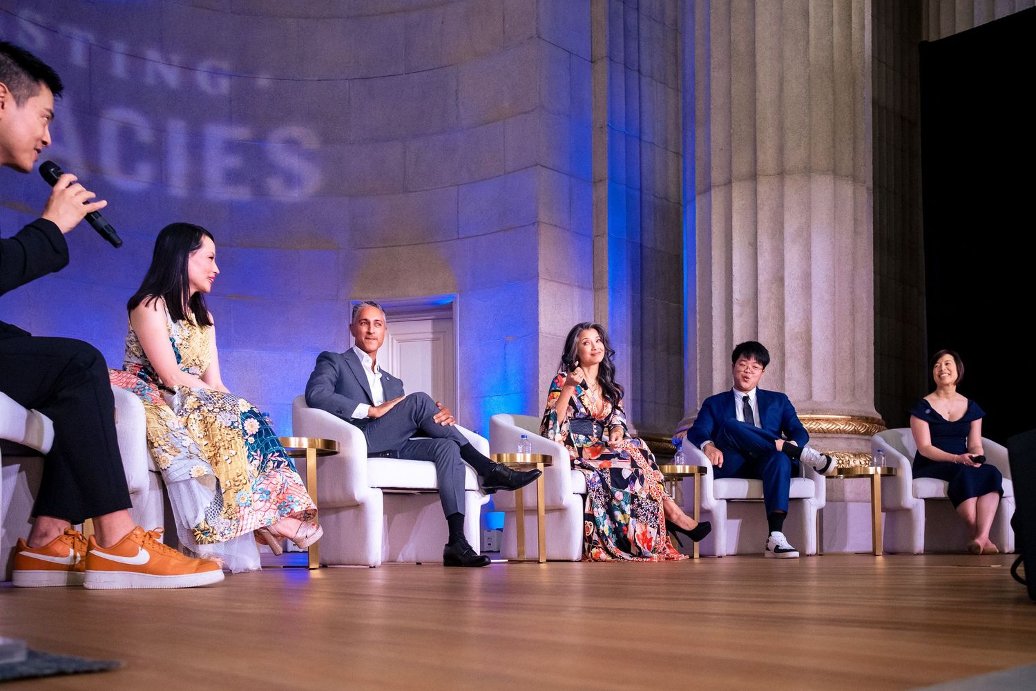 Panel discussion on stage with six speakers seated in elegant venue with classical columns and event backdrop.