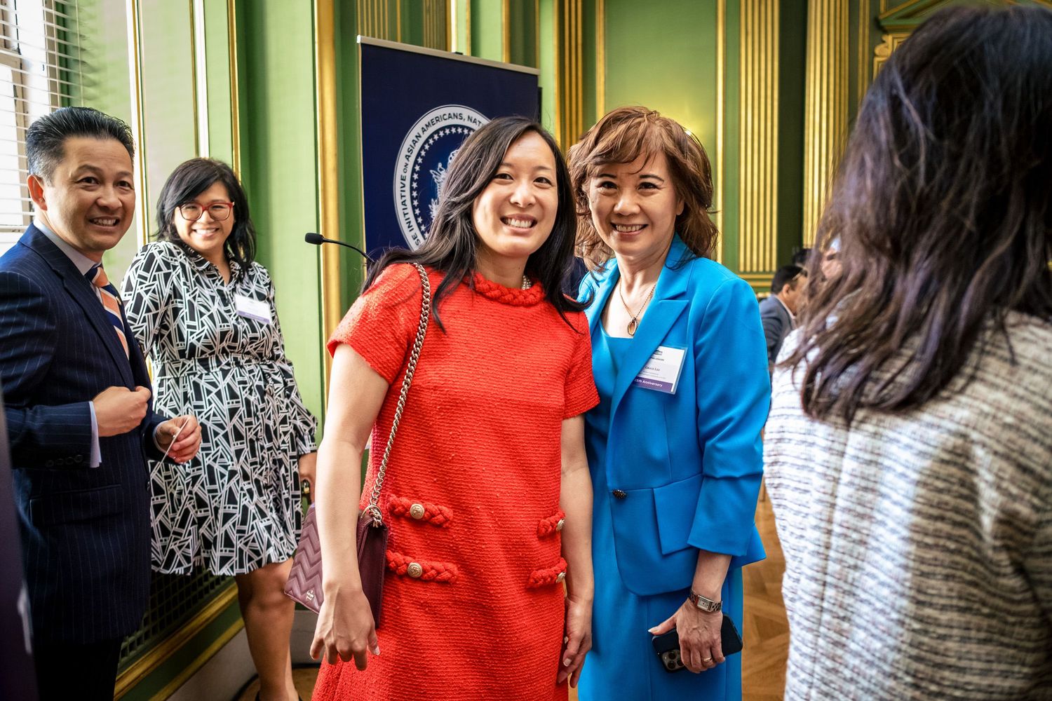 People networking at a formal event near a presidential seal banner, with women in red and blue outfits smiling.