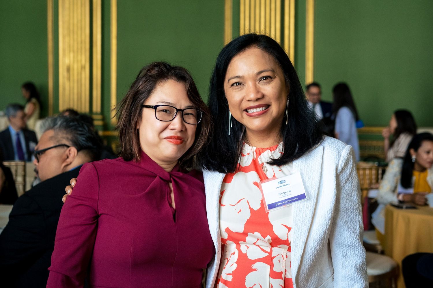 Two women smiling at a formal event in an elegant green-walled venue with guests in the background.