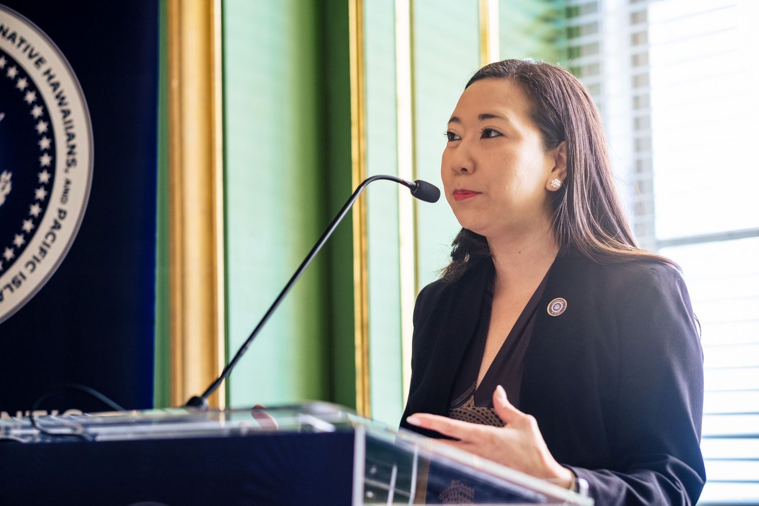 Asian woman in black blazer speaking at podium with microphone at official government event.