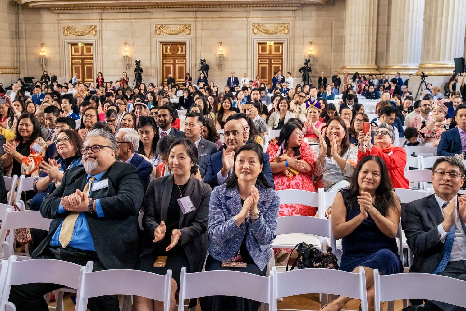 Diverse crowd of people clapping and smiling at an indoor event in a grand hall with white chairs.