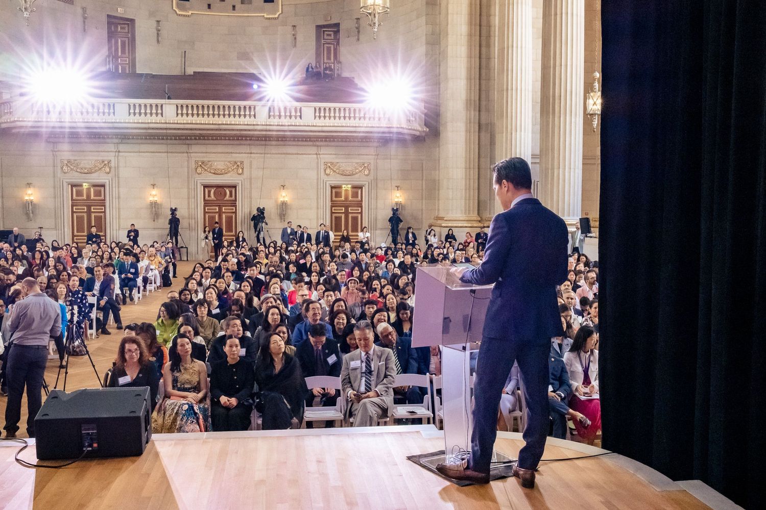 Speaker addresses large audience in grand hall with bright stage lights during a formal public event.