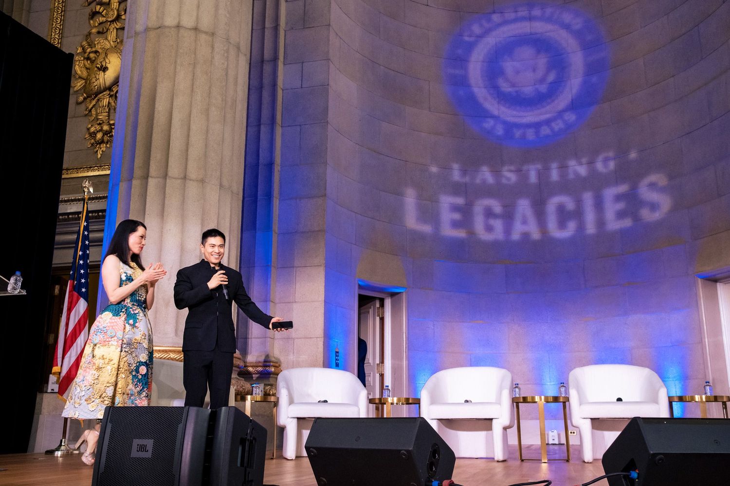 Two hosts speak on stage at a Lasting Legacies celebration event, with elegant columns and blue logo backdrop.