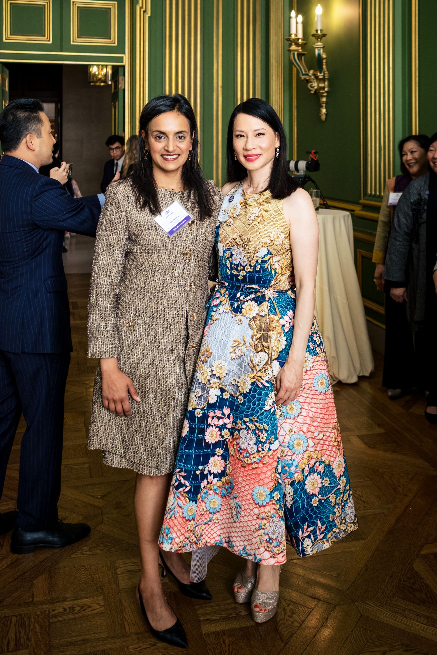 Two elegantly dressed women pose together at a formal event in a green-paneled ballroom.