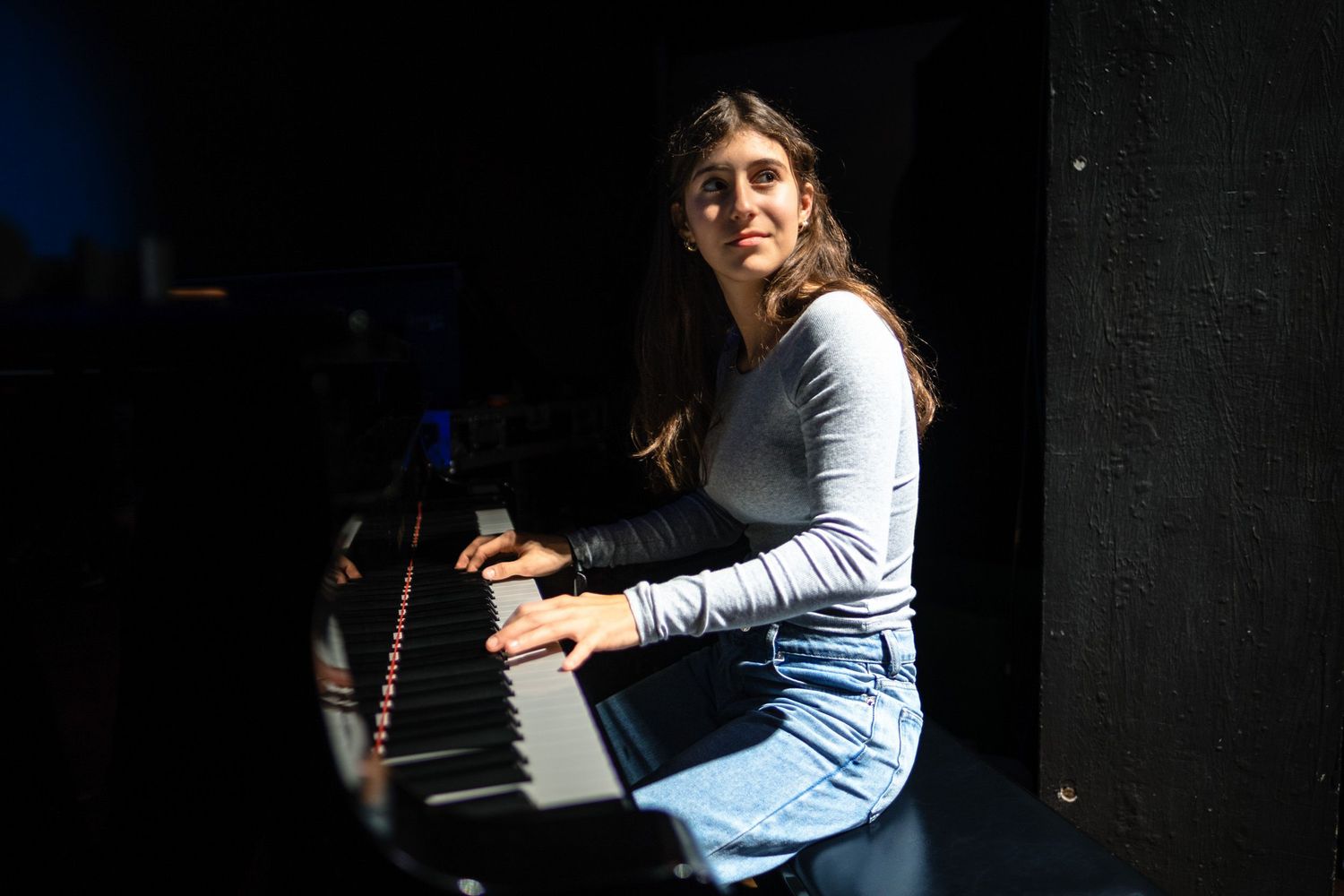 Young woman playing a black piano, smiling over her shoulder in a dramatic spotlight.