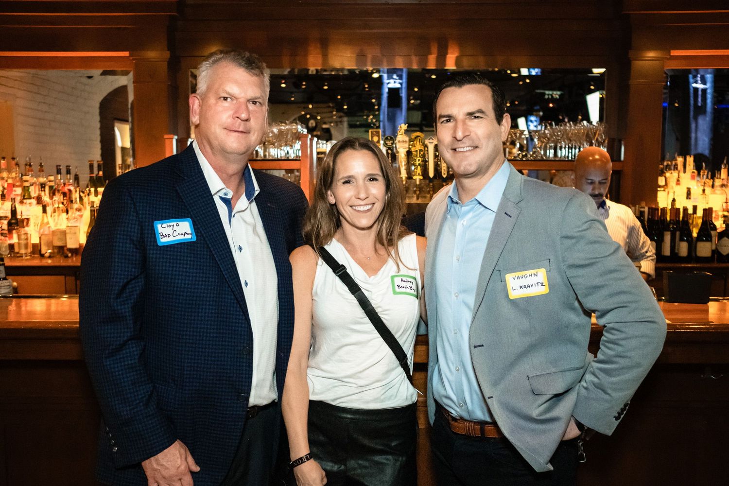 Three professionals smiling together at a bar networking event, wearing name tags, with a well-stocked bar in the background.