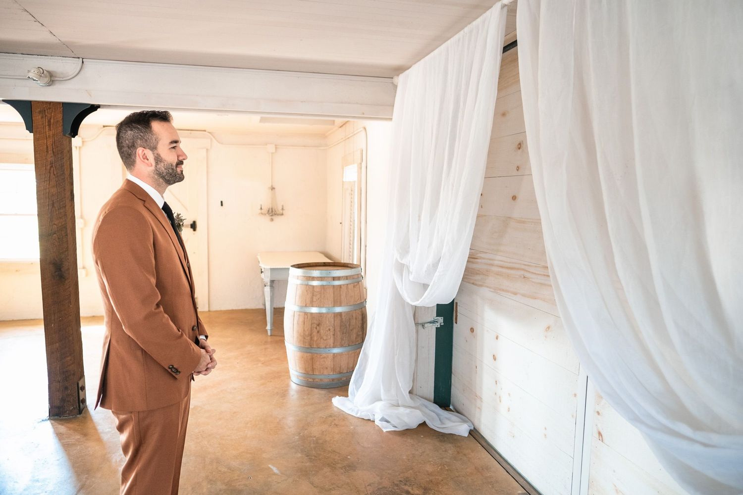 Groom in elegant brown suit standing alone in rustic wedding venue with white draping.