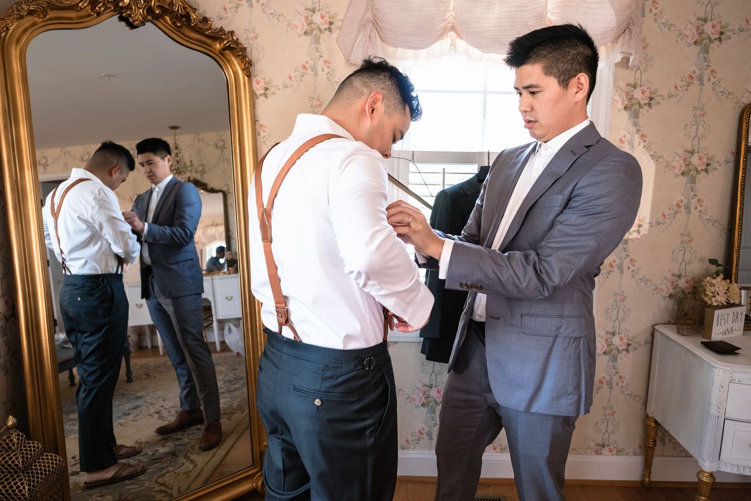 Groomsman helps groom with tie while another adjusts his suspenders before the wedding ceremony.