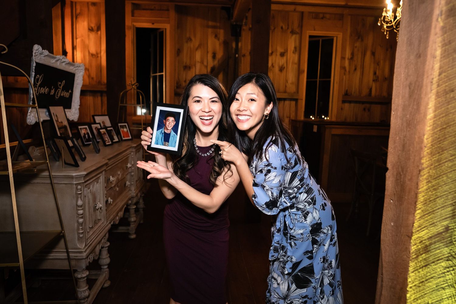 Two women smile and pose together holding a framed photo at a rustic barn wedding reception.