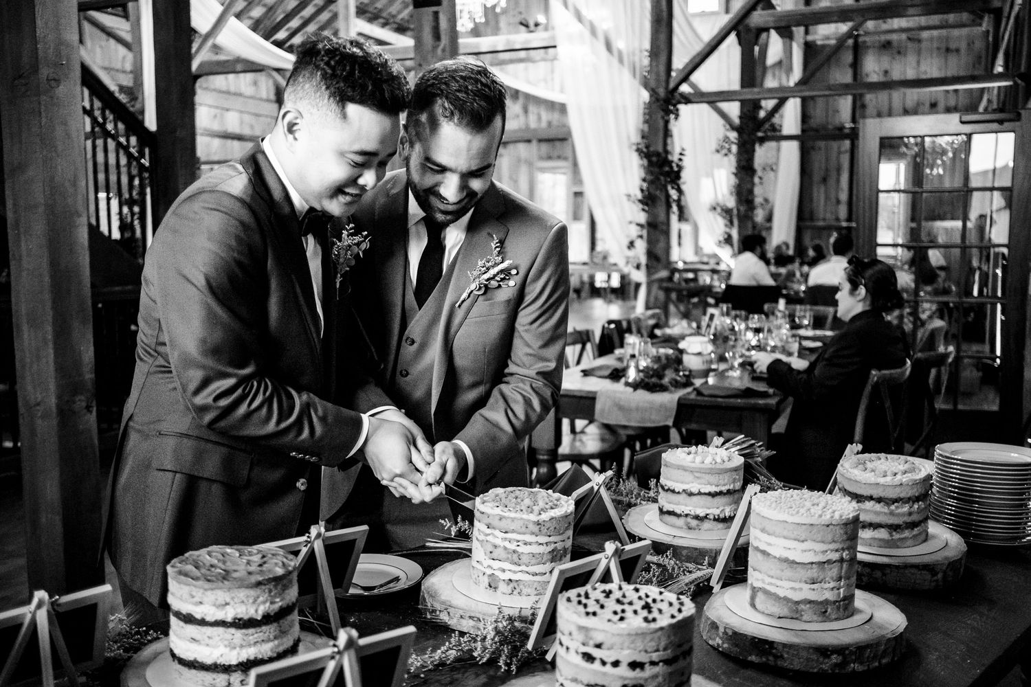 Two grooms cut wedding cake together in black and white photo at rustic barn reception.