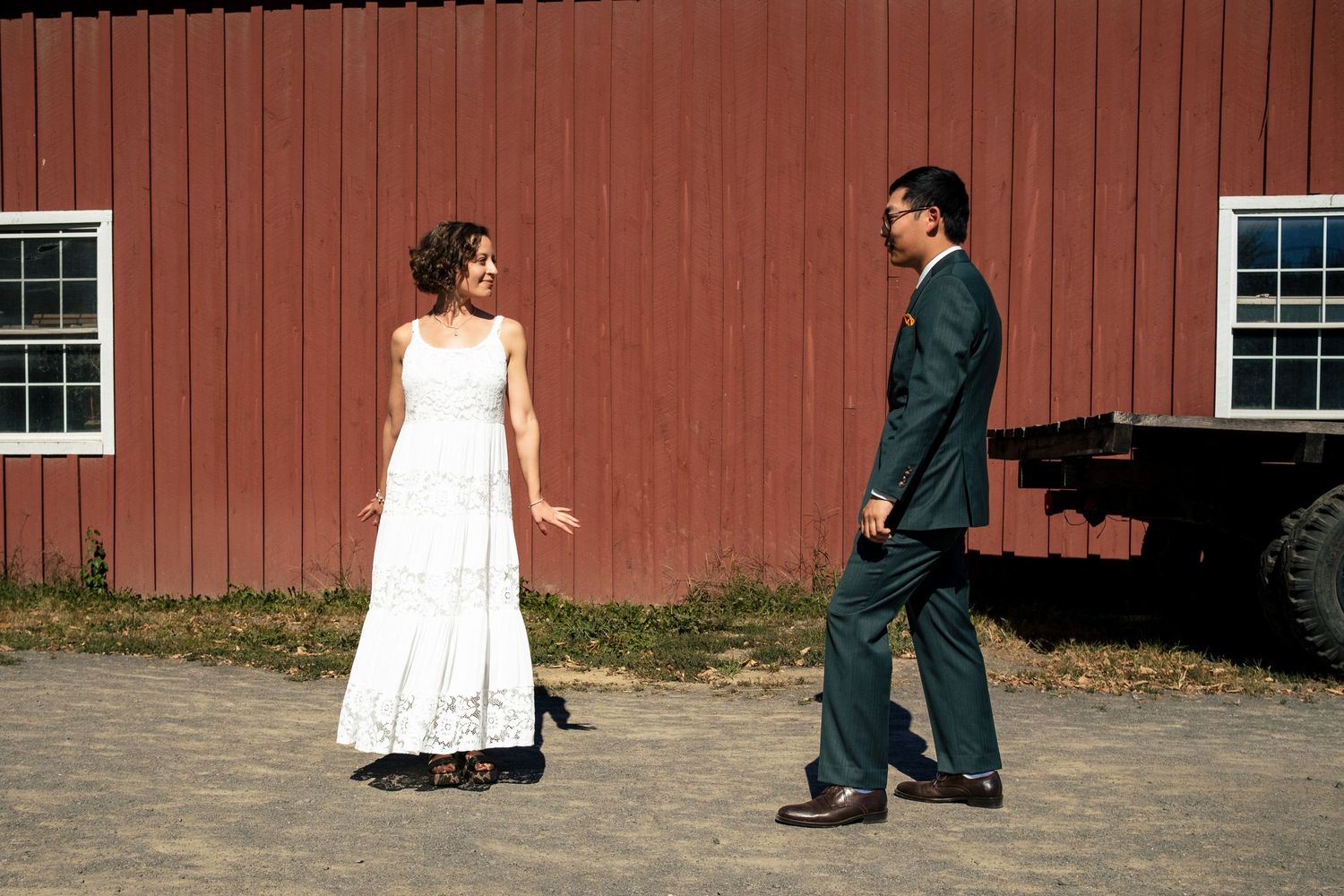A bride in white lace dress and groom in teal suit share a candid moment by a red barn.