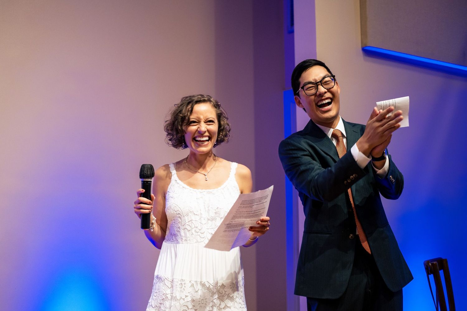 Bride and groom laughing together on stage during wedding reception speeches with microphone.