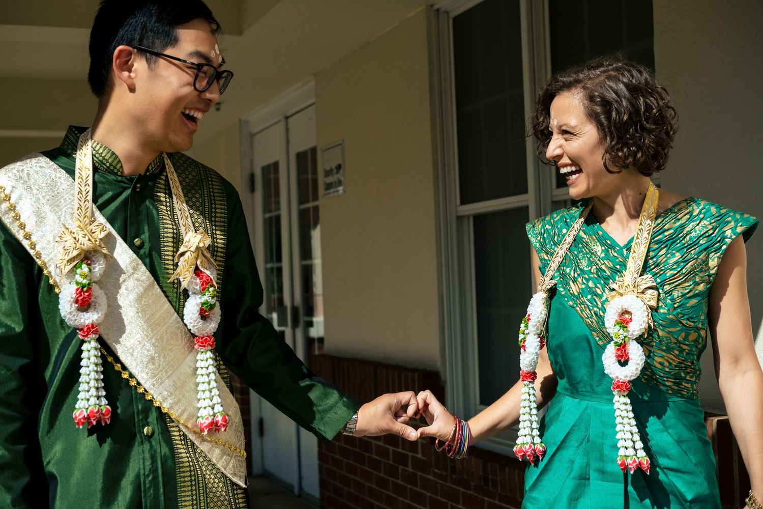 Bride and groom in traditional Thai green wedding attire laughing together on porch.