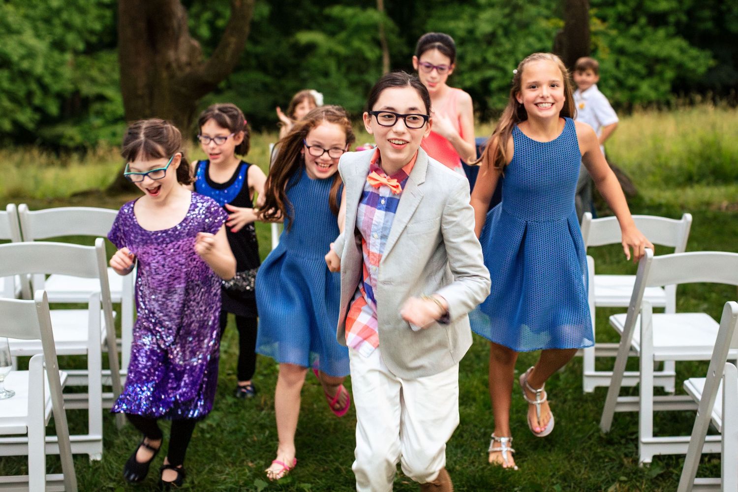 Children in formal attire running joyfully through white chairs at an outdoor wedding ceremony on a green lawn.