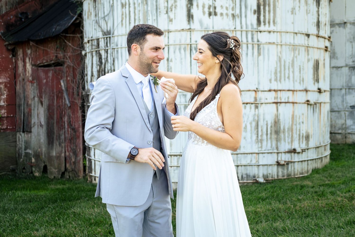 Bride adjusts grooms boutonniere while smiling together in front of rustic weathered grain silo.