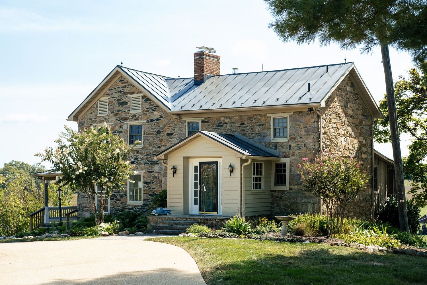 Charming stone farmhouse with metal roof, yellow trim, and lush landscaping on a sunny day.