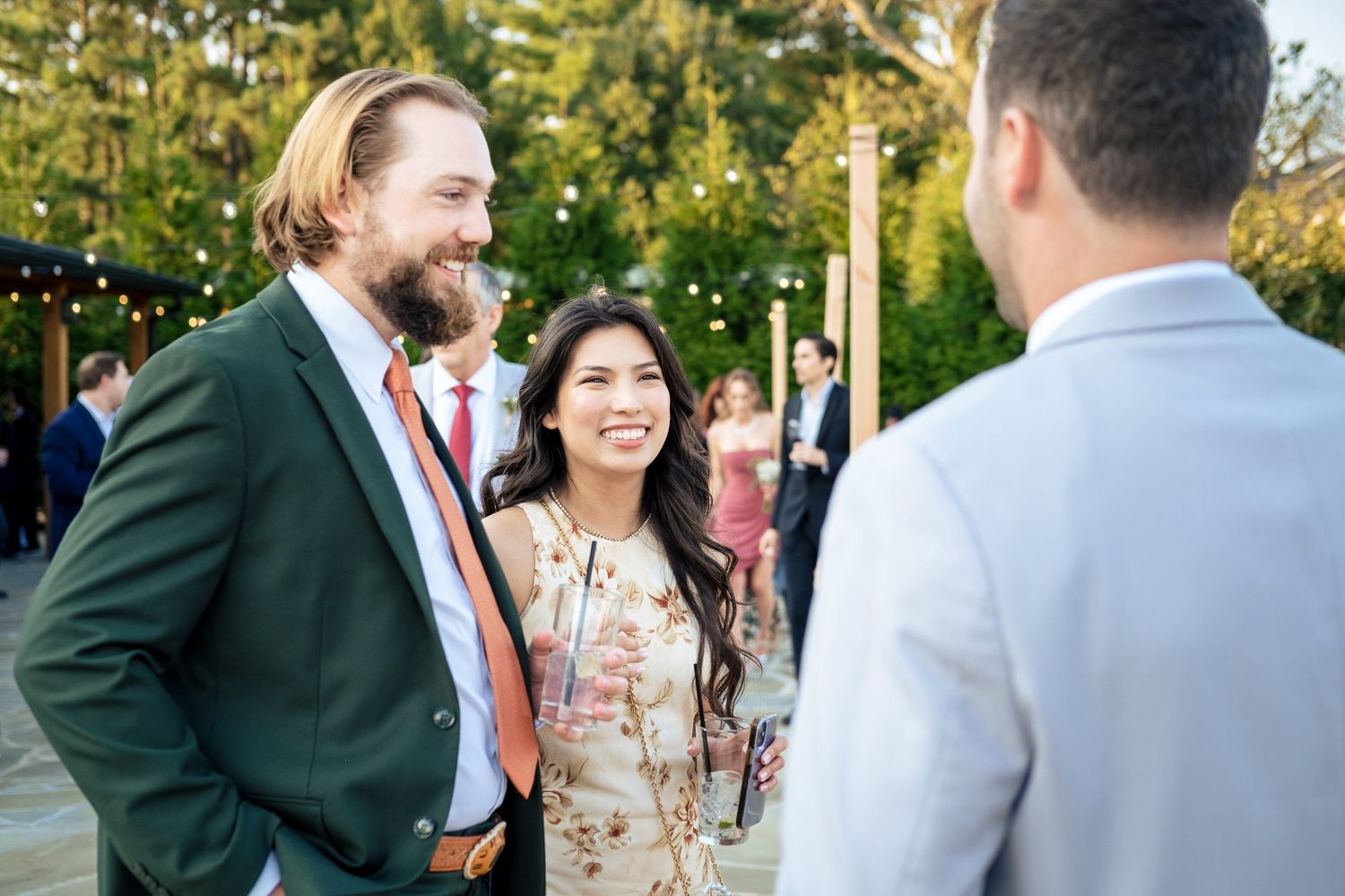 Wedding guests in formal attire smile and converse outdoors at a reception with string lights in background.