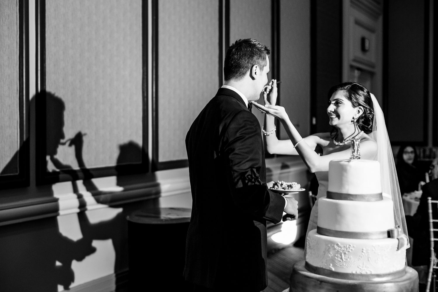 Bride playfully smears wedding cake on grooms face beside a white tiered cake.