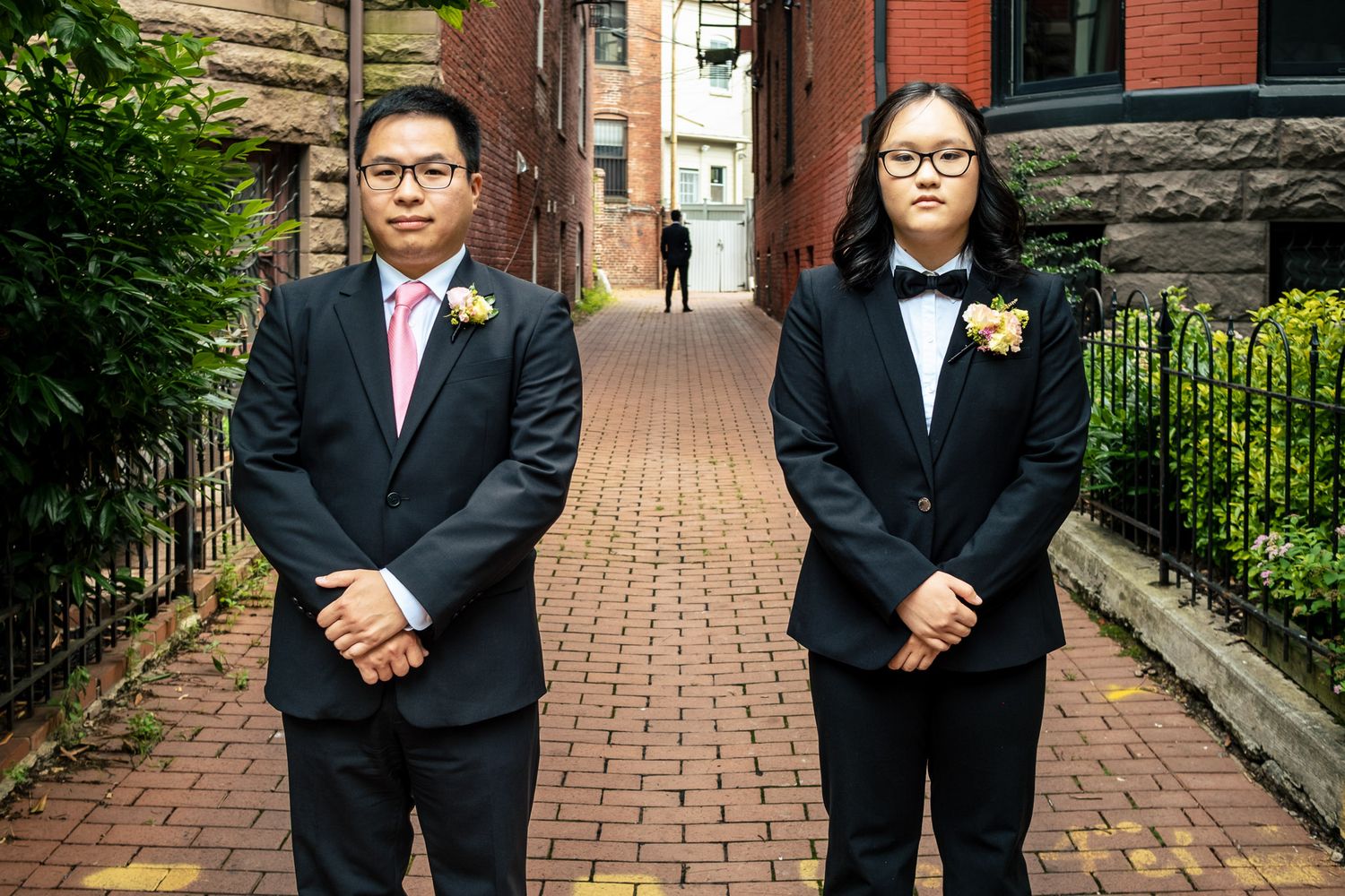 Two wedding attendants in black suits with floral boutonnieres stand on a brick pathway.