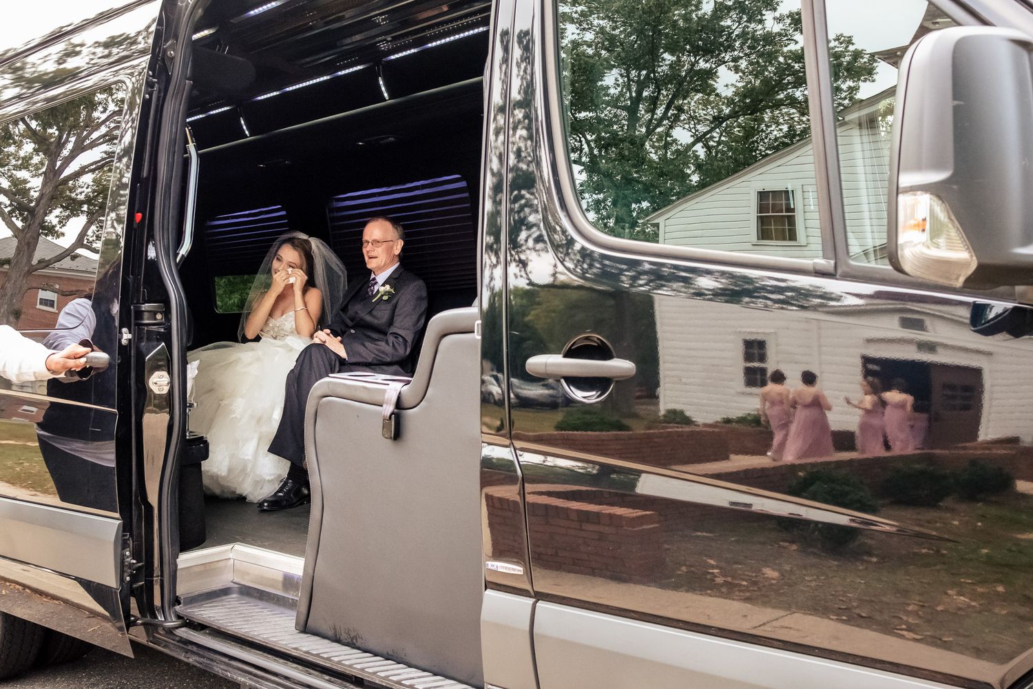Bride and groom sit in luxury black wedding limo with bridesmaids reflected in door.