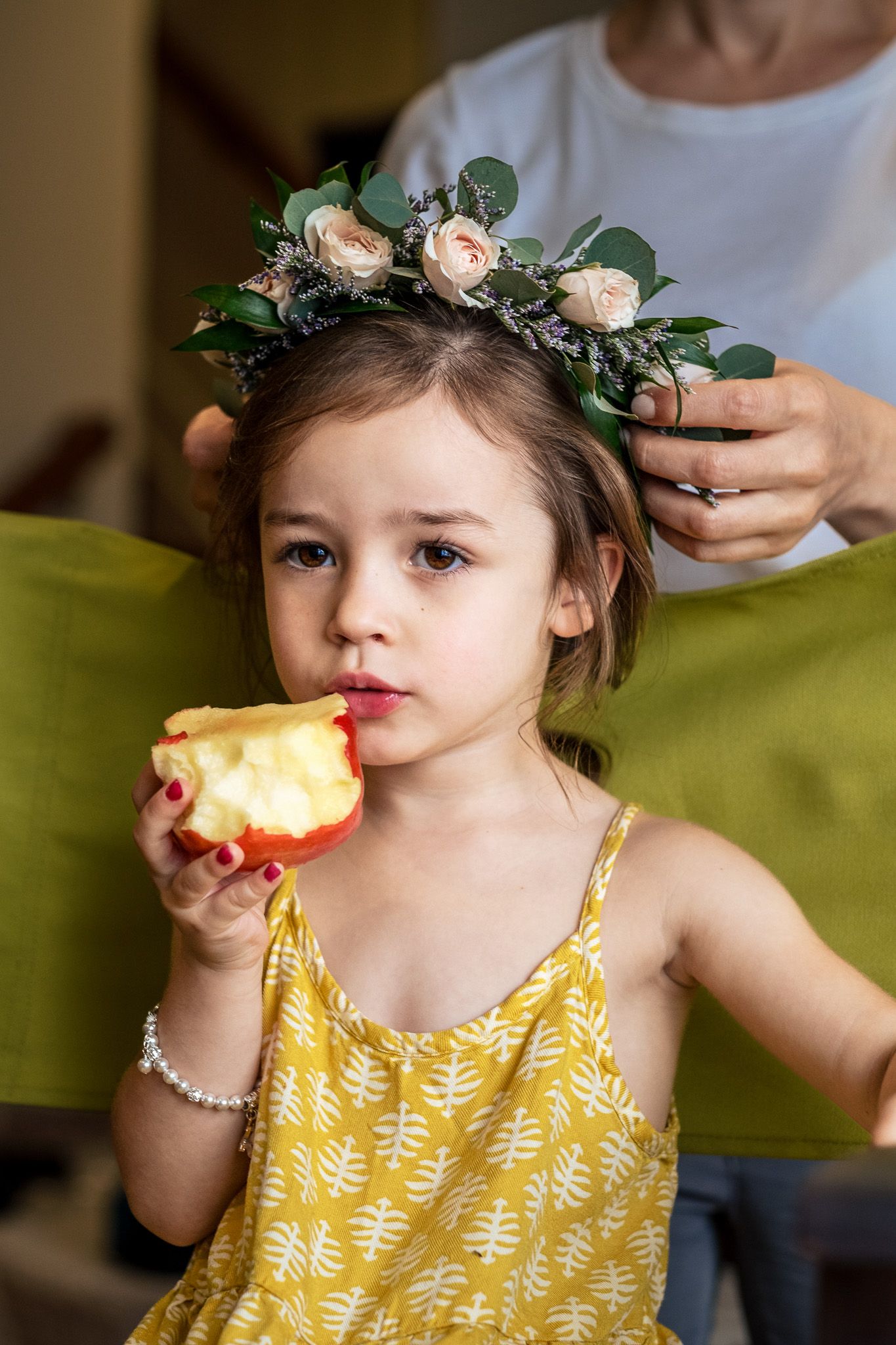 Adorable flower girl in yellow dress and floral crown eats an apple during wedding preparations.