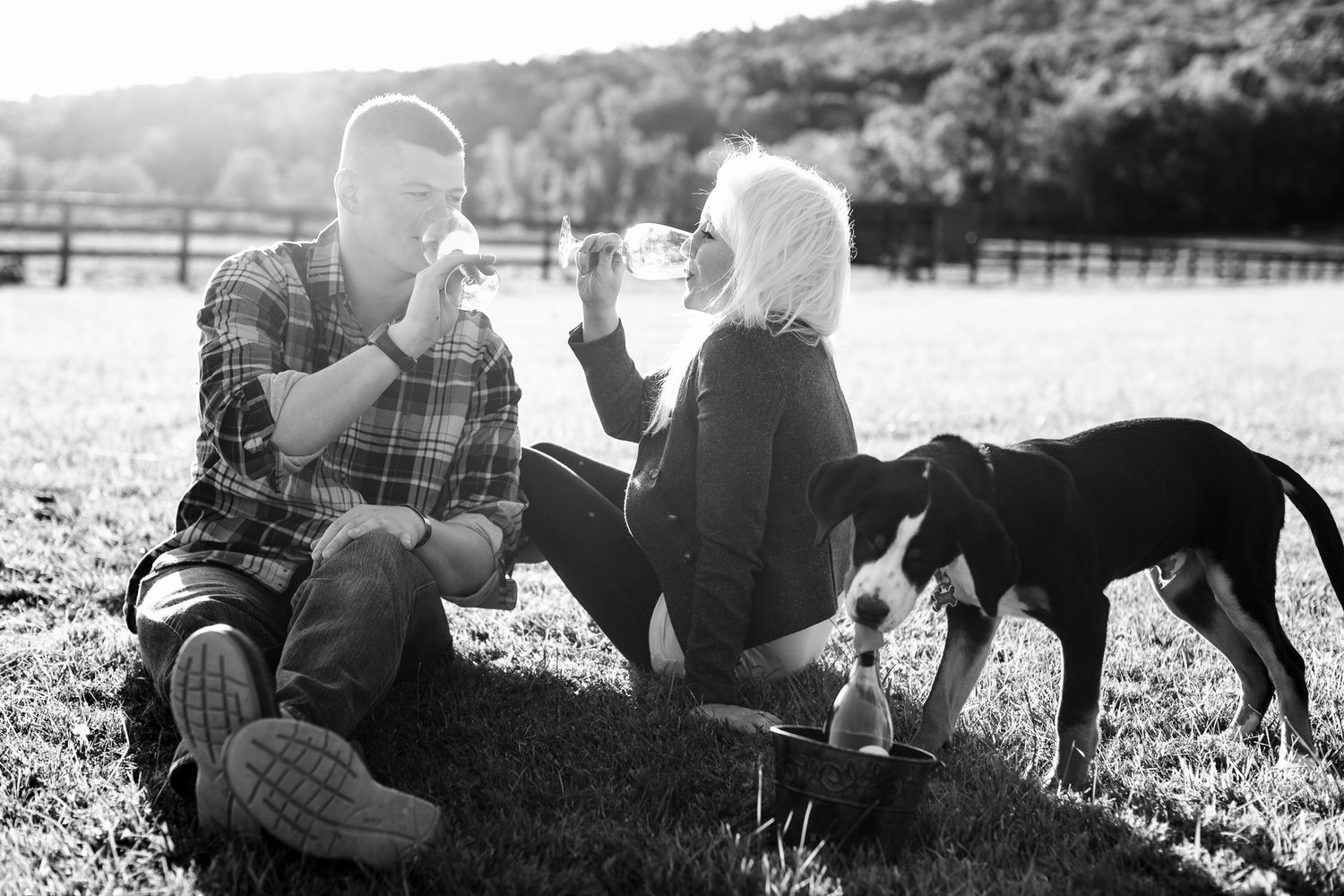 Couple sips wine outdoors on grass with their dog in black and white photo.