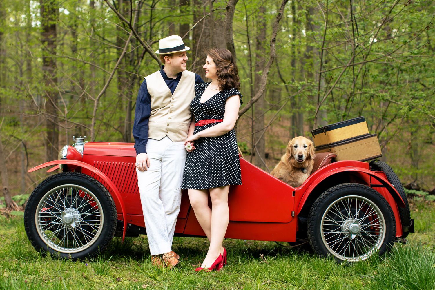 Vintage-dressed couple standing by a classic red convertible with a golden retriever in a forest.