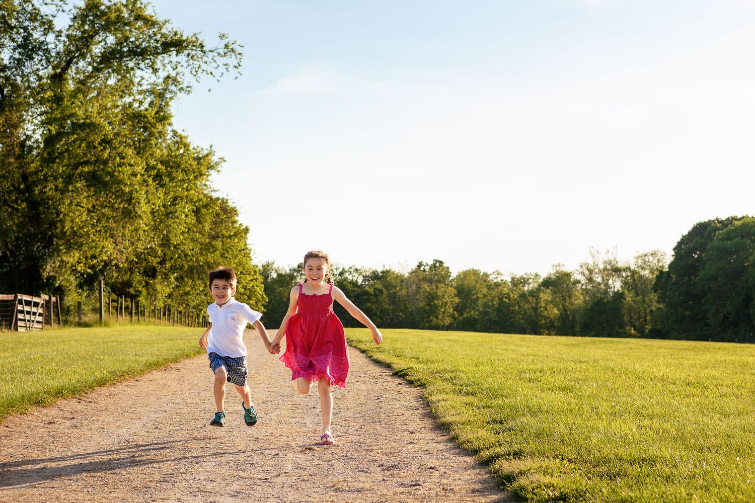 Two young children running hand-in-hand down a sunlit gravel path surrounded by green fields.
