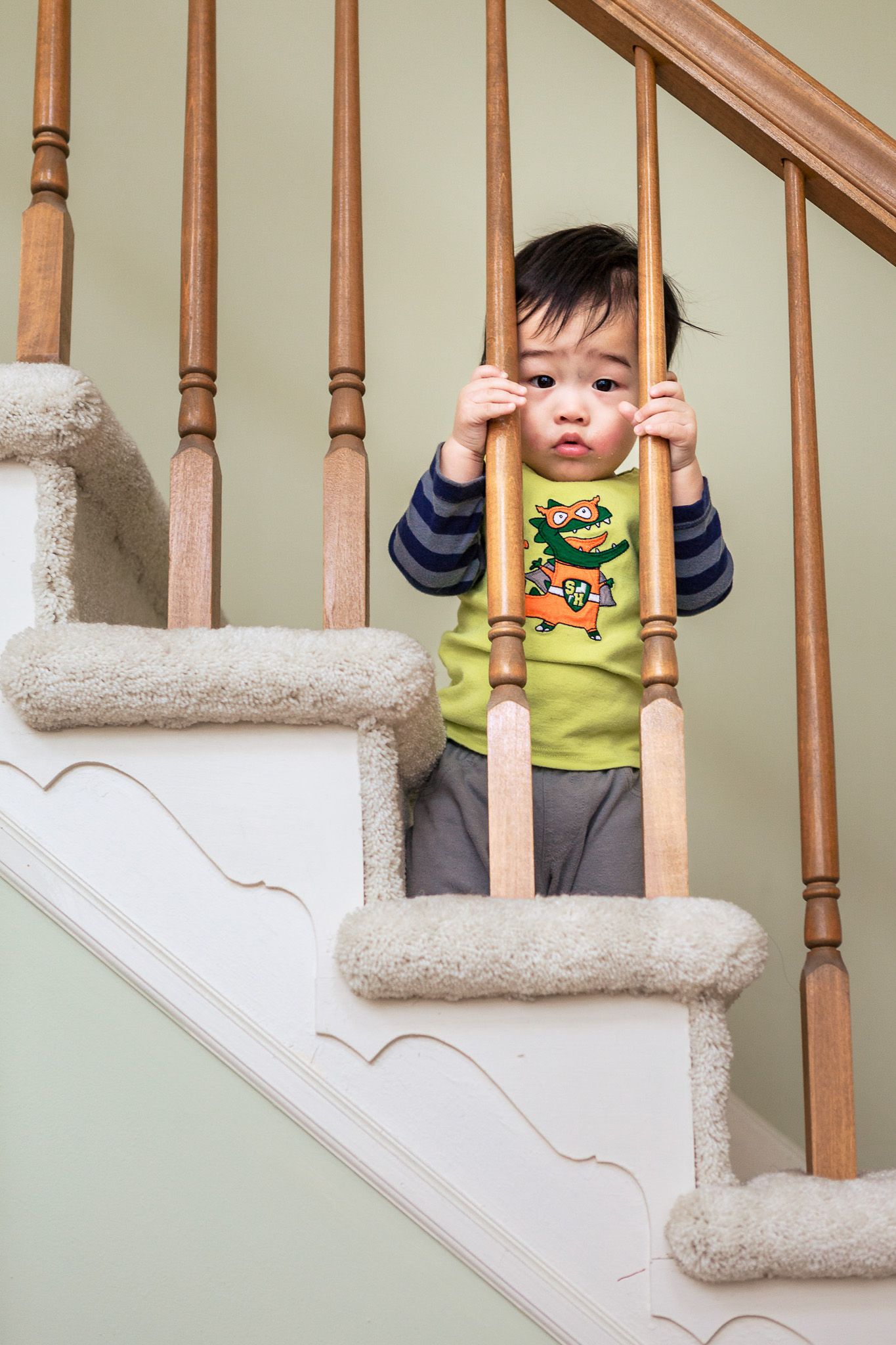 Curious toddler peering through wooden stair railings on carpeted home staircase.