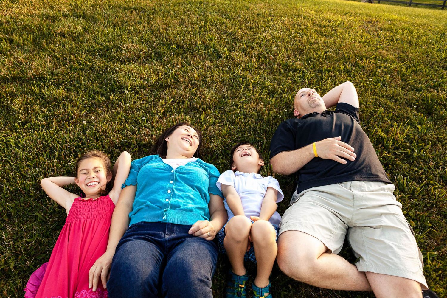 Family of four lying on green grass laughing together on a sunny day in the park.