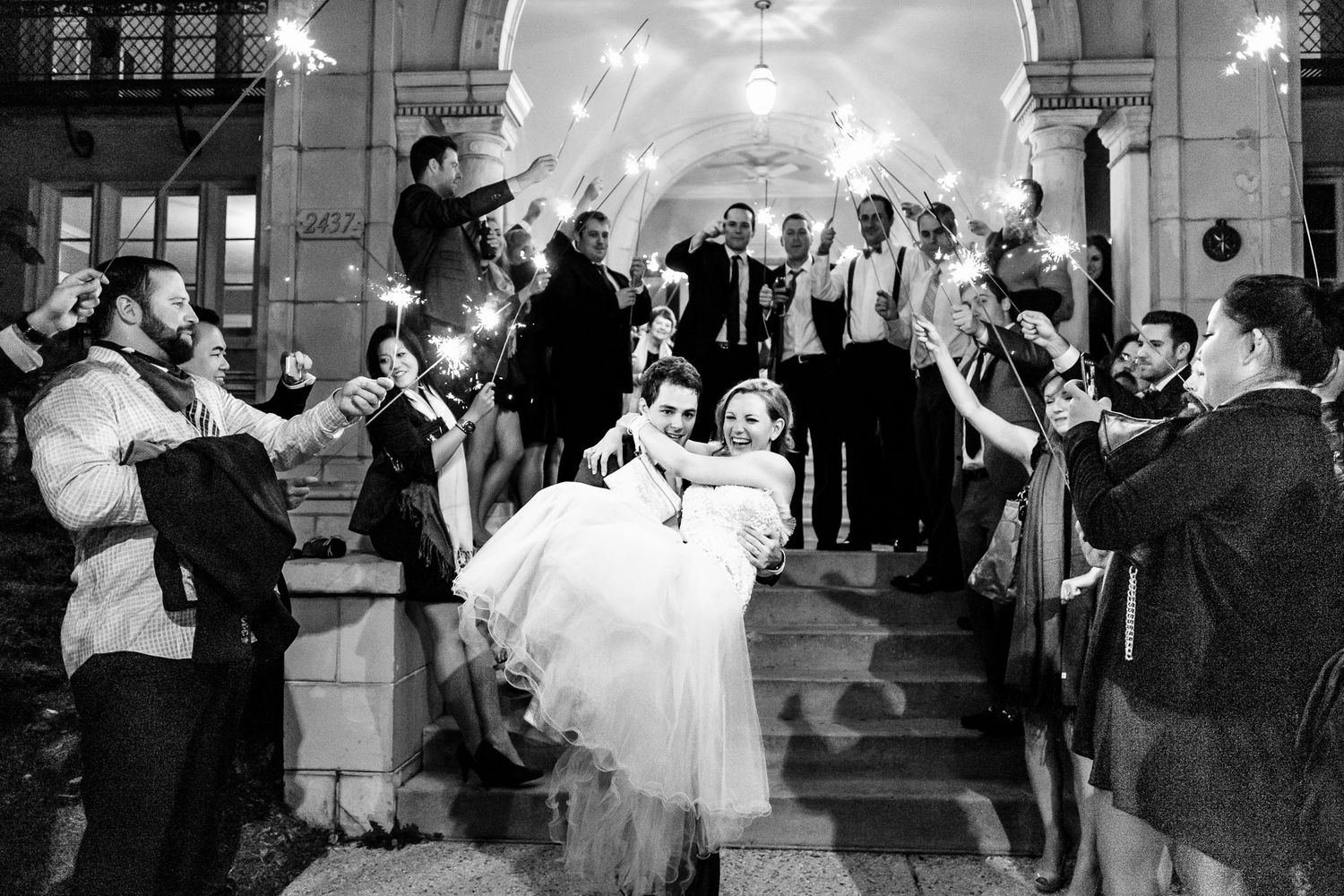Groom carries bride through sparkler exit surrounded by cheering guests at night wedding.