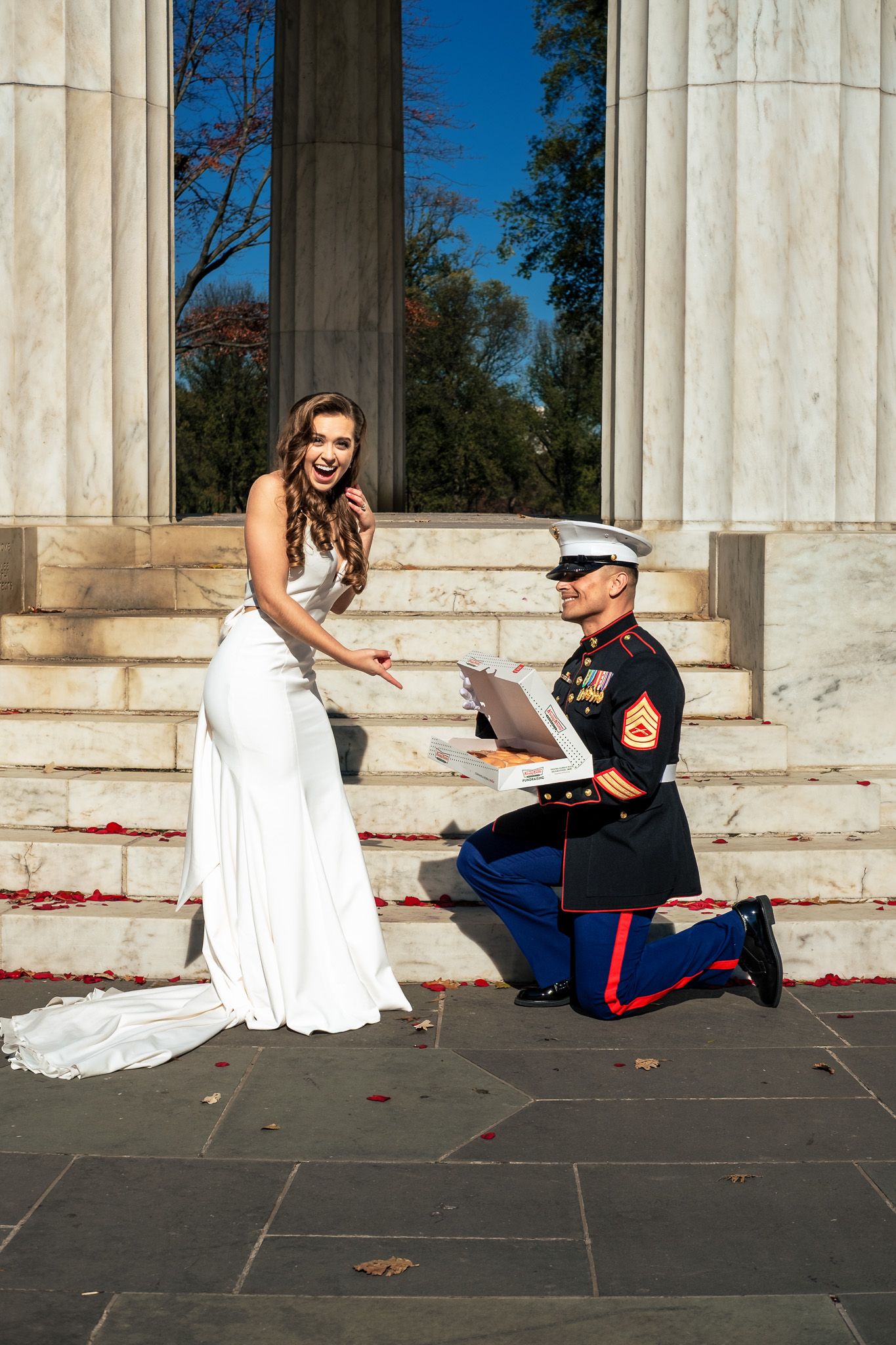 Marine in dress uniform kneeling before smiling bride on monument steps in autumn setting.