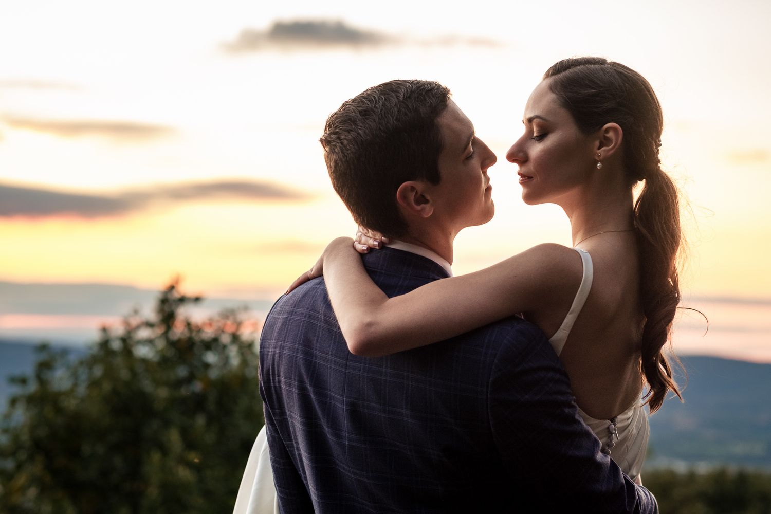Bride and groom share a romantic embrace at sunset with a colorful sky backdrop.